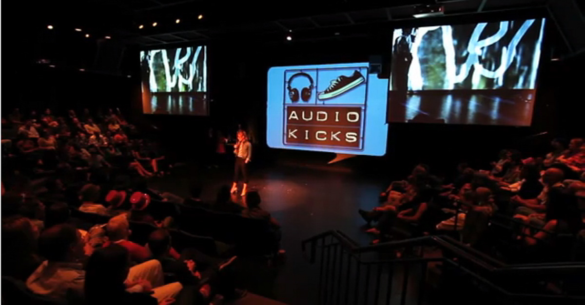 Live performance of Audio Kicks on stage, with a title screen reading Audio Kicks and an audience seated in the theater