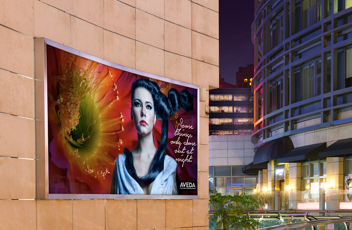 Aveda concept billboard mockup featuring a model with dark upswept hair against a large flower, shown installed on a city building at night
