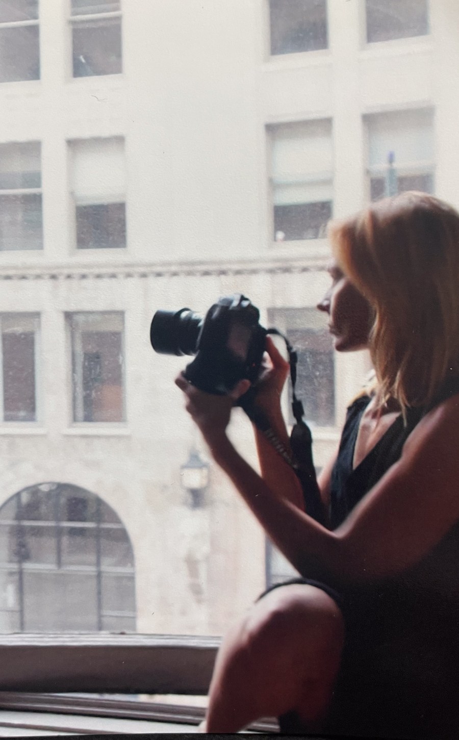 Photographer Renae Vespremi at a window with a camera, a cityscape behind her